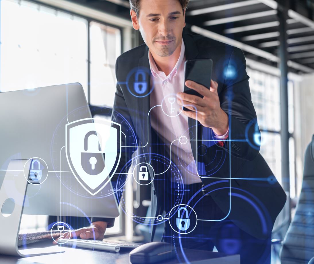 Männliche Person mit Handy vor Laptop. Im Vordergrund schweben Sicherheitsschlösser.